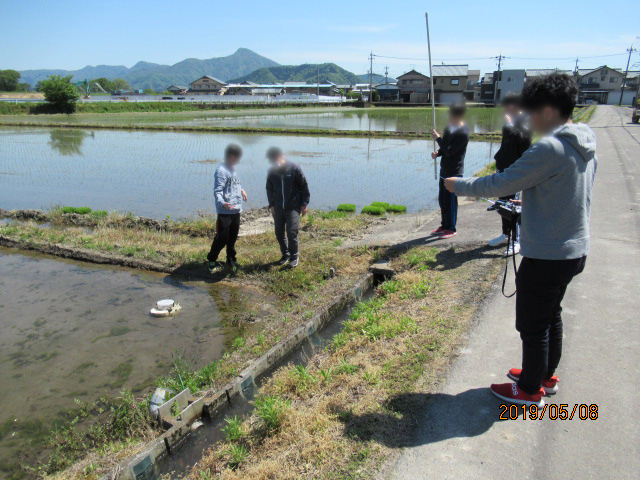 良く晴れた青空のもと、水が張ってある水田の周りで学生ら5名が小型ロボットの実験をしている様子